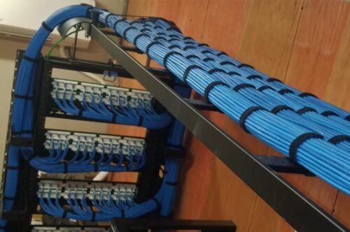 A neatly organized network cable management system with blue cables is displayed on racks, resembling a professional computer repair workspace. The cables are bundled along a black metal framework on a wooden floor, showcasing a tidy setup. computer repair