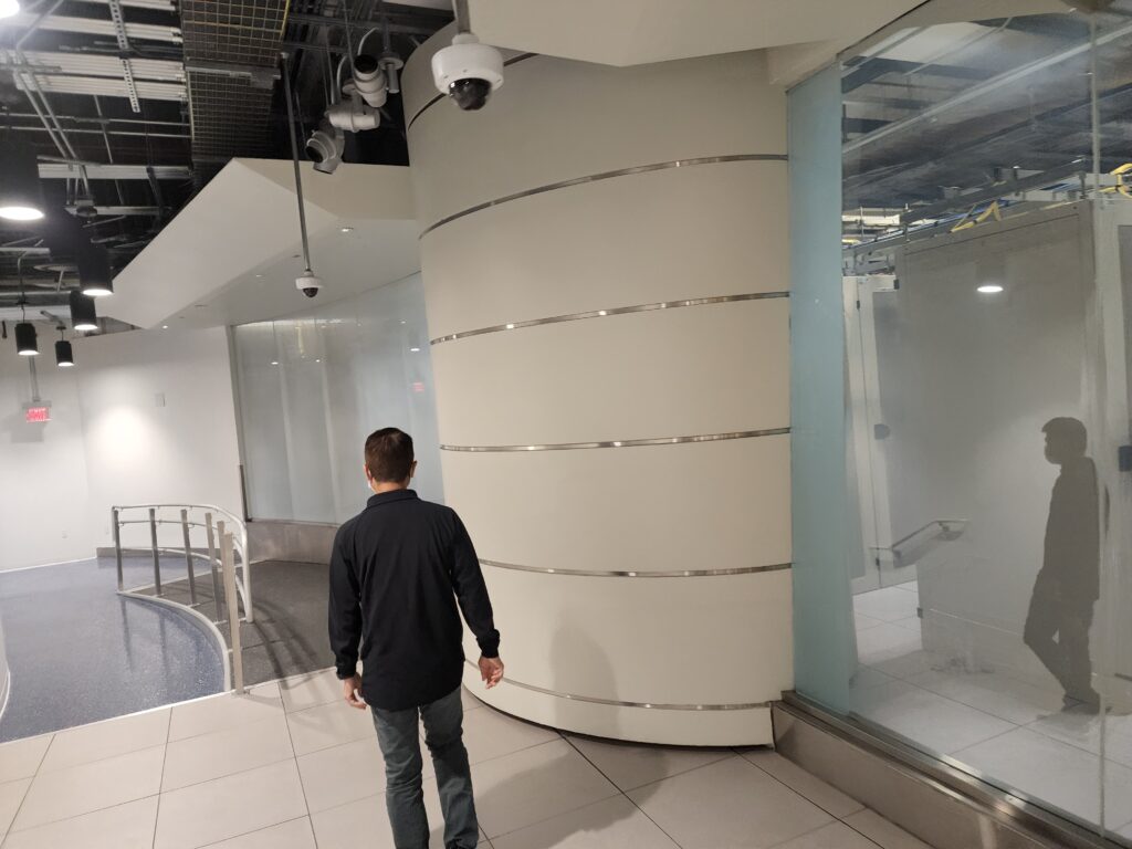 A person strolls through a modern indoor space with white walls and tiled floors, resembling a sleek computer repair shop. The ceiling is partially exposed, revealing pipes and ducts. A glass wall lines the right side, while a security camera keeps watch from above. computer repair