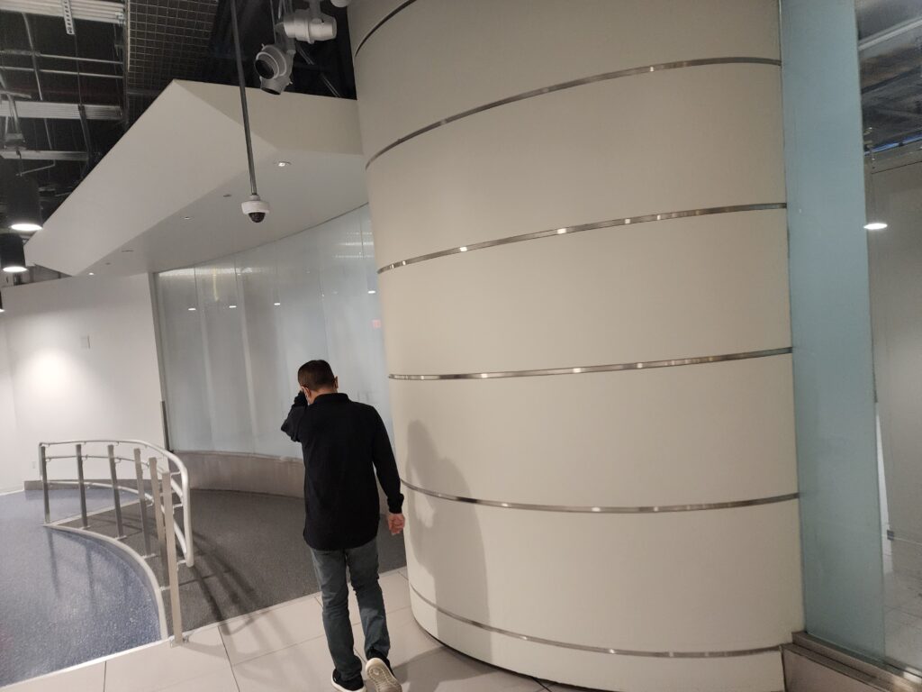 A person in a black jacket and jeans walks through a modern, minimalistic interior reminiscent of a sleek computer repair shop. The space features curved walls with metal accents, large glass panels, and overhead lighting, creating an airy and spacious atmosphere. computer repair