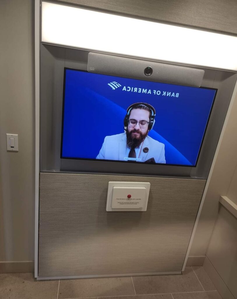 A wall-mounted video screen shows a bearded man wearing headphones, discussing computer repair. The backdrop features the Bank of America logo. Below the screen, a white panel with a button is prominently displayed. computer repair