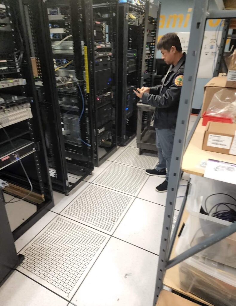 A person in a server room stands checking a smartphone, amidst the hum of computer repair. The room is filled with server racks containing various electronic equipment and cables, while shelves with additional tools for repairs are visible on the right. computer repair