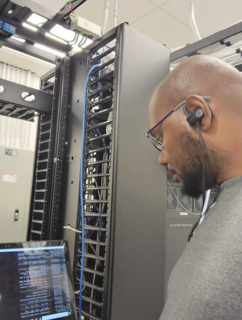 A person wearing glasses and earbuds is intently working on computer repair in a server room, surrounded by equipment racks. They are focused on a screen displaying code or data, with cables and electronic equipment visible around them. computer repair