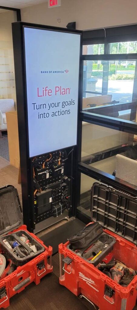 Digital display under maintenance in an office building, showing a screen with Bank of America Life Plan: Turn your goals into actions. Open toolboxes with various tools and computer repair equipment are scattered on the floor nearby. computer repair