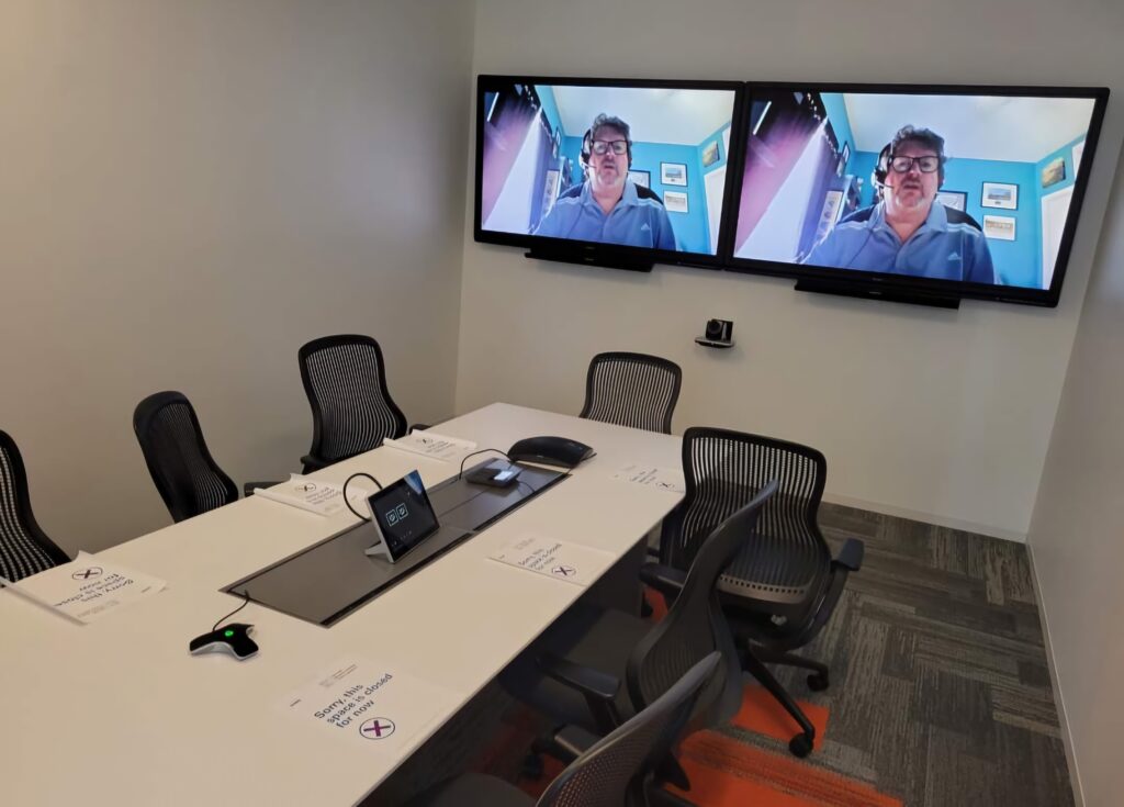 The conference room features a long table, office chairs, and two screens showing a person on a video call. Notices emphasize social distancing. A remote control and conference phone are on the table, alongside materials related to computer repair for troubleshooting sessions during meetings. computer repair
