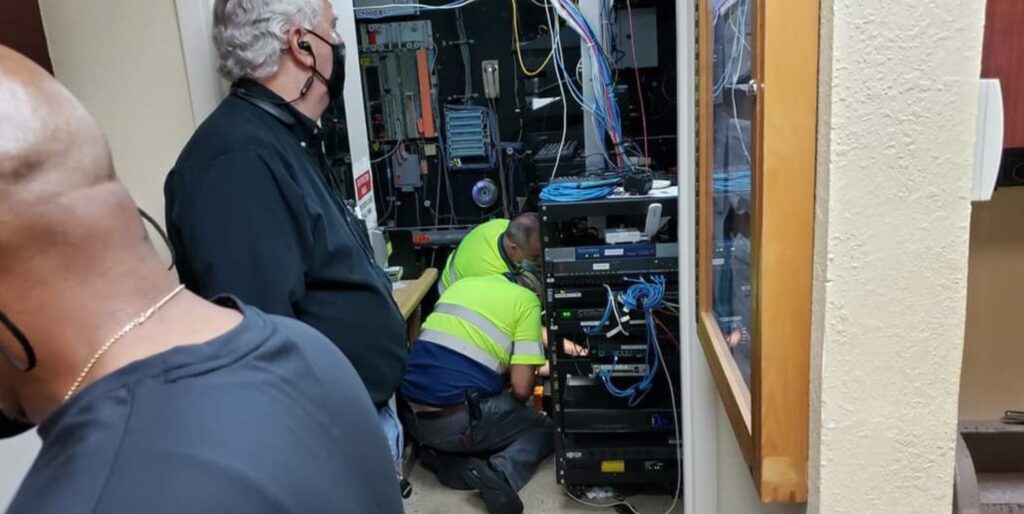 Two individuals, one wearing a bright yellow vest, are working on a server rack with multiple cables in a small room. Another person stands nearby observing the computer repair tasks. They appear to be addressing technical or IT maintenance tasks. computer repair