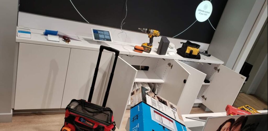 A room with open cabinets and various tools on a counter, including a drill and tool bags, sits ready for computer repair. A boxed item is on the floor, and a black screen on the wall displays white text. computer repair