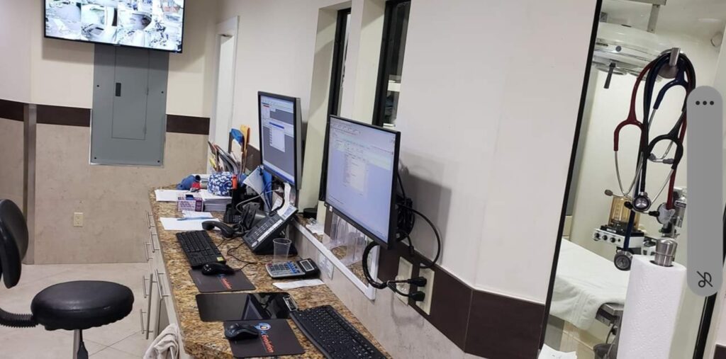 A veterinary clinic reception area with two computer monitors on a granite counter, hinting at recent computer repair. Various medical equipment, including a stethoscope, hangs nearby. A wall-mounted screen displays multiple surveillance camera feeds in the background. computer repair