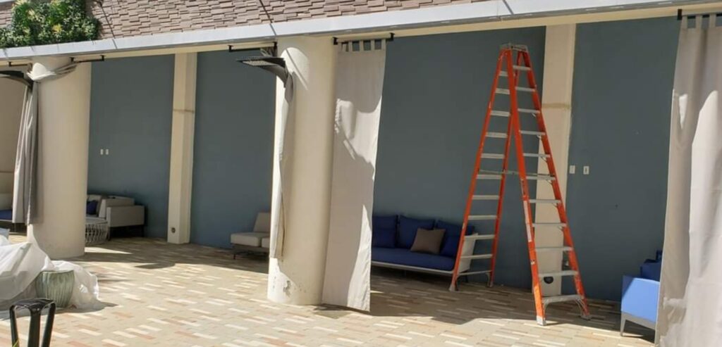 A patio with an orange ladder leans against a wall under a shaded area, perfect for contemplating computer repair challenges. White curtains hang from the ceiling, dividing the space. Blue cushioned seating lines the wall, while sunlight casts playful shadows on the brick floor. computer repair