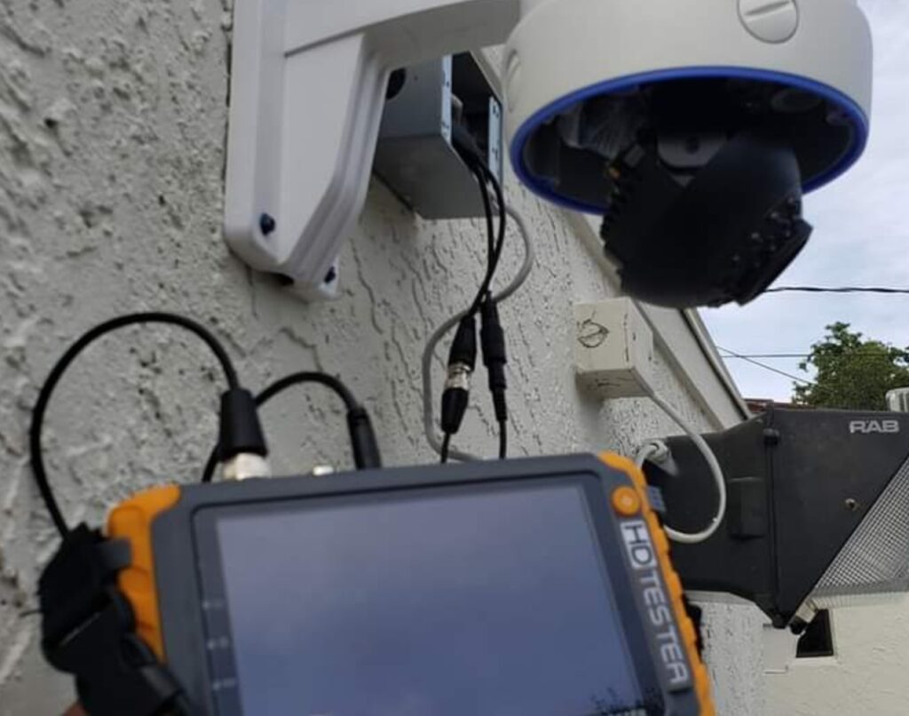 A person is using an HD tester to install or configure a surveillance camera mounted on a textured wall. Cables connect the tester to the camera, with another camera visible in the background.