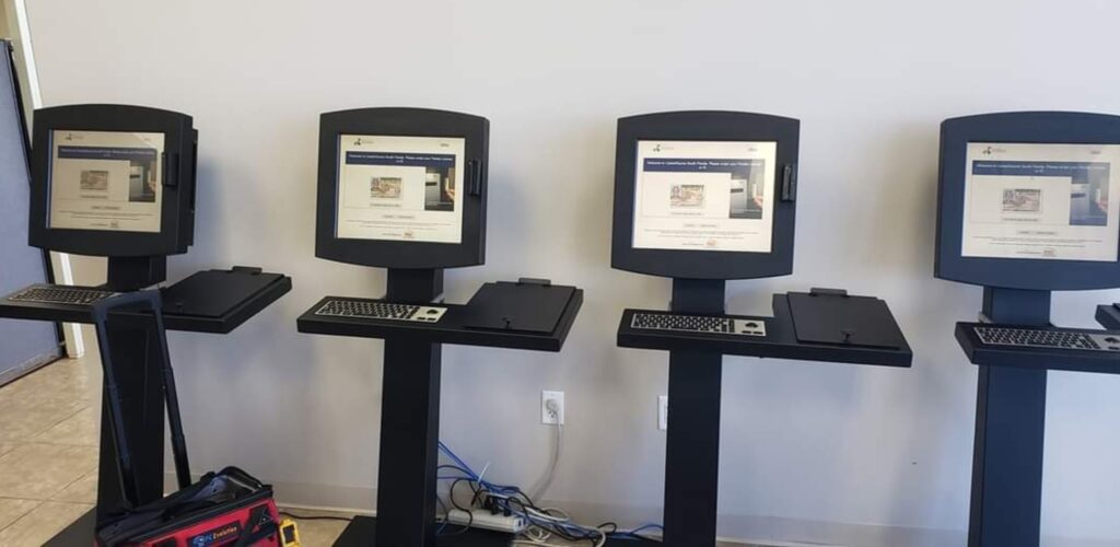 Four voting machines stand in a row, each with a black screen and keyboard. Cables and equipment are visible on the floor. The machines are set up in a plain room with light-colored walls.