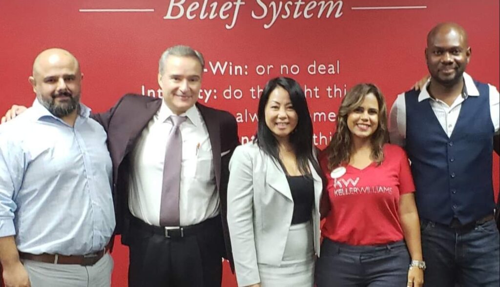 Five people stand smiling in front of a red wall with text about a belief system. They are dressed in business attire, with two men in suits and three individuals in casual professional wear.