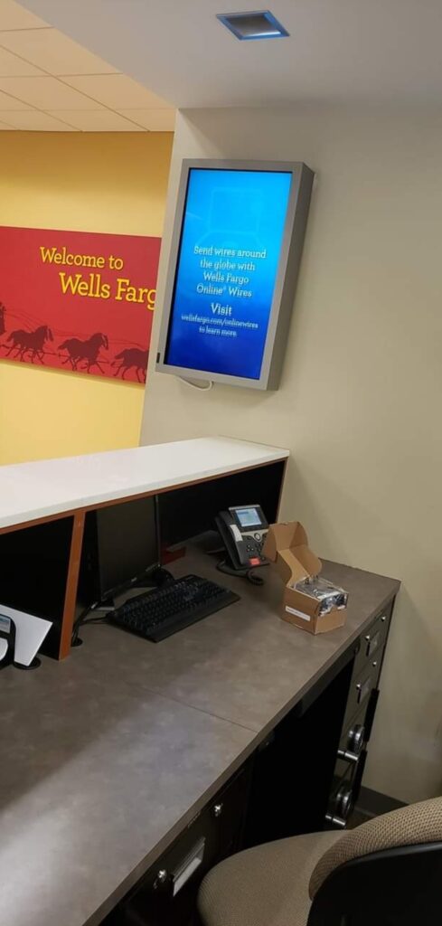 A bank office with a desk featuring a computer, phone, and open cardboard box. In the background, a wall displays a Wells Fargo sign and a digital screen promoting online wire transfers.