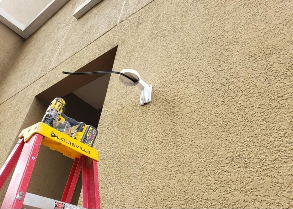 A red ladder stands against a textured beige wall, with a power drill and a case labeled Louisville resting on top. A white security camera is mounted on the wall, with a black cable extending from it.